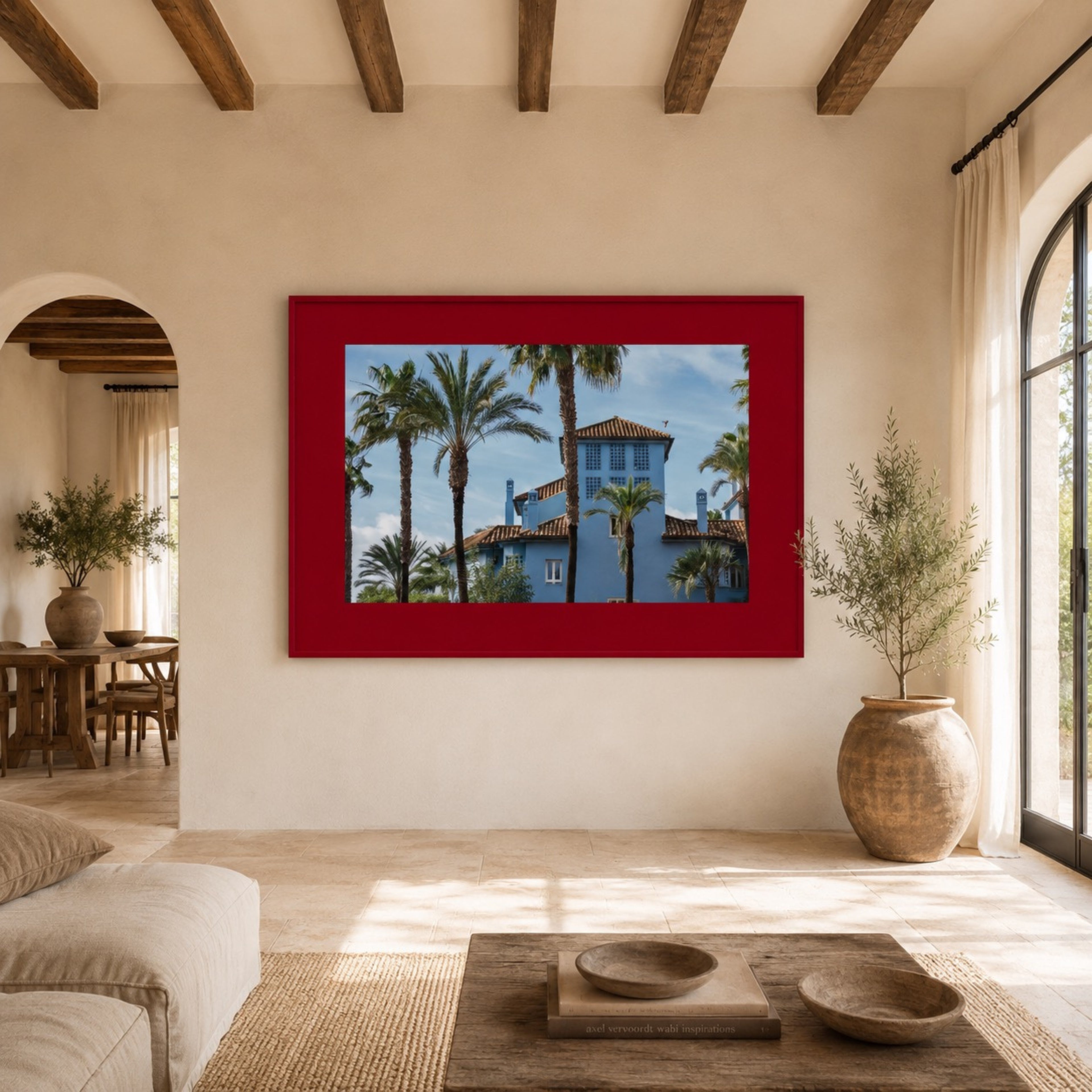 A framed art print depicting a blue Mediterranean-style villa surrounded by palm trees, set in a bright, modern room with a red frame.