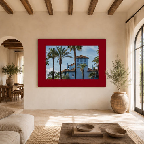 A framed art print depicting a blue Mediterranean-style villa surrounded by palm trees, set in a bright, modern room with a red frame.